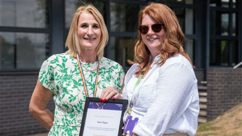 Two people standing in front of a building with large windows; one holding a framed certificate